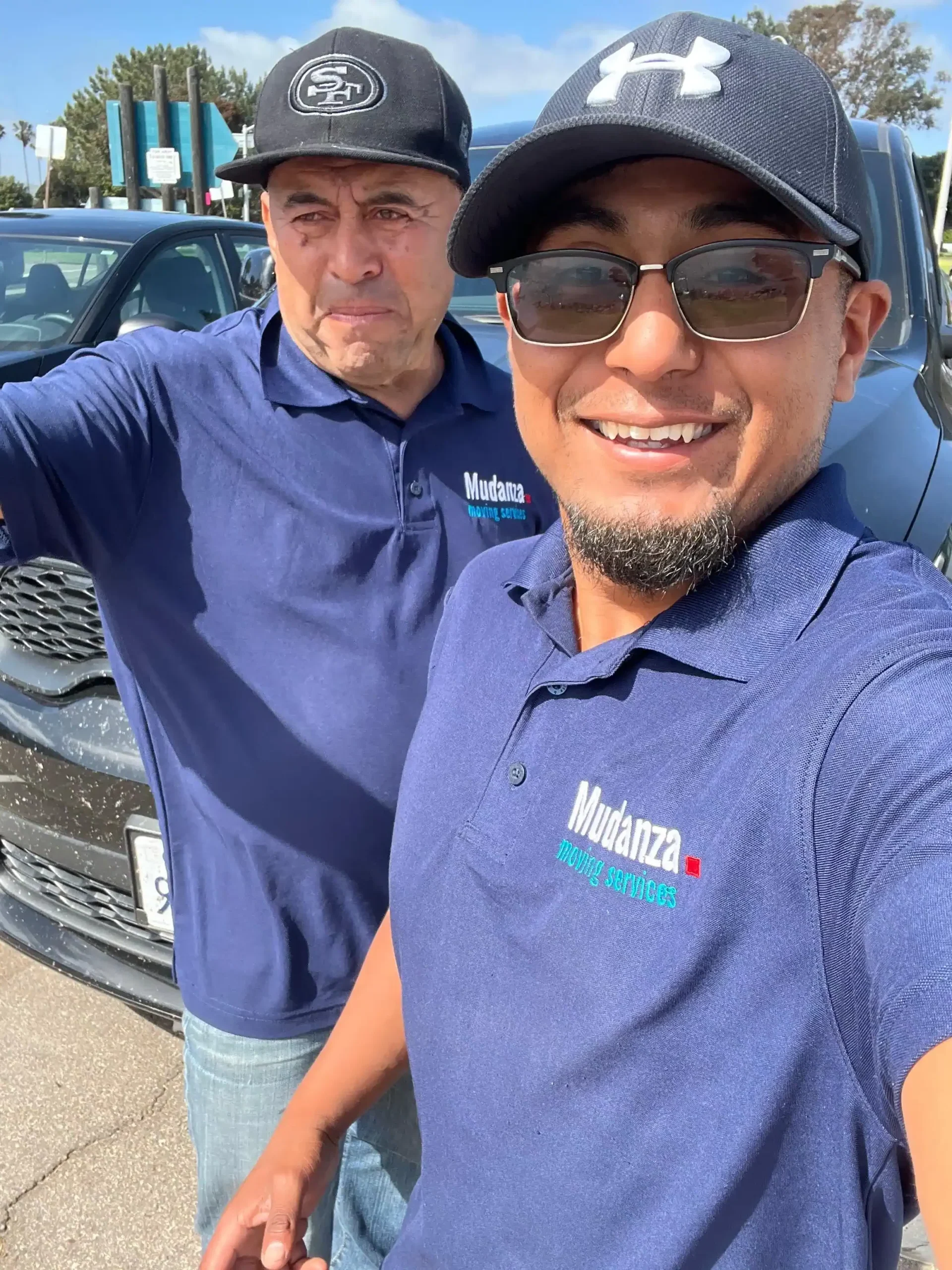 Two smiling professional movers wearing branded Mudanza Moving Services shirts standing in front of a dark moving truck on a sunny day.