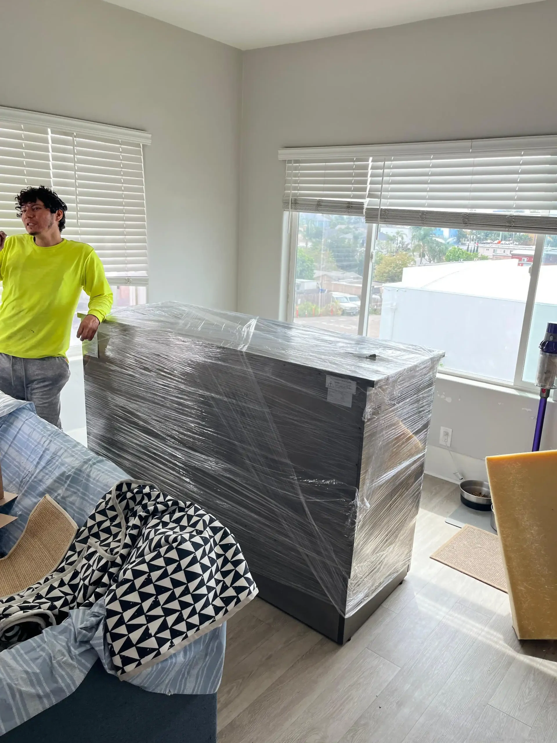 A large, heavy piece of furniture completely protected in layers of plastic shrink wrap next to a Mudanza Moving Services crew member.